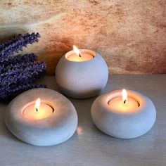 three white candles sitting on top of a table next to some lavender flowers and a purple plant