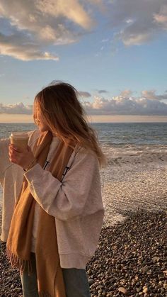 a woman standing on top of a rocky beach next to the ocean holding a coffee cup