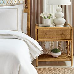 a bed with white sheets and pillows next to a lamp on a nightstand in front of a window