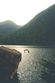 a person jumping into the water from a rock in front of some hills and trees