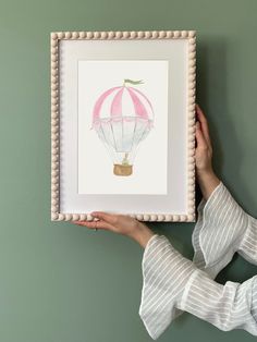 a woman holding up a framed painting with a hot air balloon on it's side