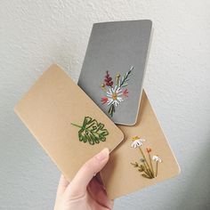 two notebooks with embroidered flowers on them are held up by someone's hand