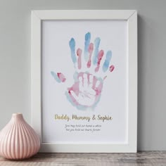 a pink and blue hand print on a white frame next to a vase with a pink flower