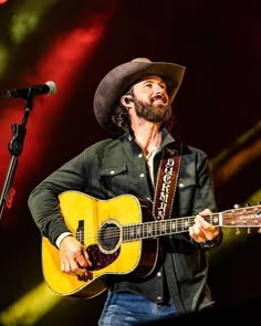 a man with a cowboy hat playing an acoustic guitar in front of a microphone on stage