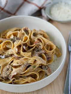 a white bowl filled with pasta covered in mushrooms and parmesan cheese next to silverware