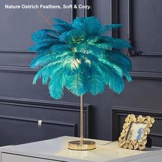 a blue feathered plant on a white table next to a gold frame and framed photograph