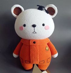 a teddy bear with an orange sweater and black hat on it's head is standing in front of a gray background