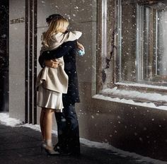 a man and woman standing next to each other in front of a building with snow falling on the ground