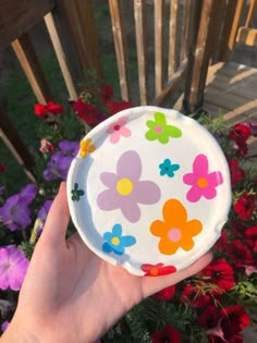 a hand holding a paper plate with flowers painted on it in front of red and purple flowers