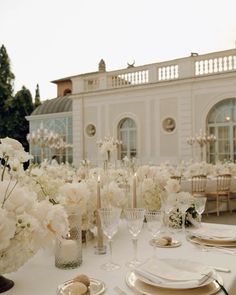 the table is set with white flowers and place settings in front of a large building