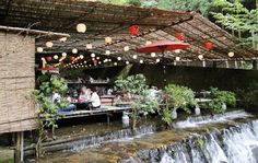 people are sitting at tables in front of a waterfall with lights hanging from the roof