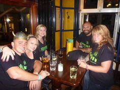 a group of people sitting around a wooden table with beers in front of them and smiling at the camera