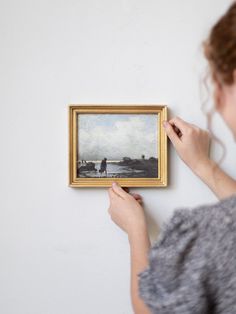 a woman holding up a painting in front of her face and looking at it on the wall