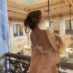 a woman in a pink dress standing on a balcony looking at the ceiling and chandelier