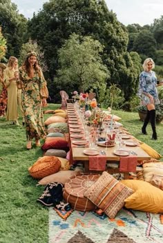 a group of women standing around a long table covered in pillows and blankets on top of a lush green field