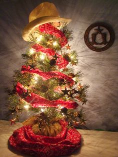 a small christmas tree decorated with red ribbon and lights in a corner next to a cowboy hat