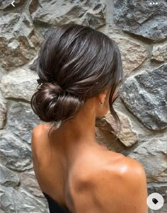 a woman with her hair in a low bun, standing against a stone wall and looking off to the side