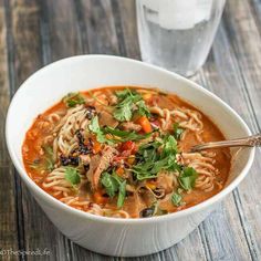 a bowl of soup with noodles, meat and veggies on the table next to a glass of water