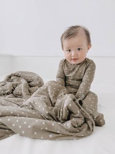 a baby sitting on top of a bed covered in a gray polka dot blanket and looking at the camera