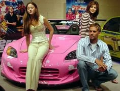 three people sitting on the hood of a pink sports car in front of a movie poster