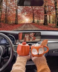 someone is holding two mugs in their hand while driving down the road with trees and leaves on both sides