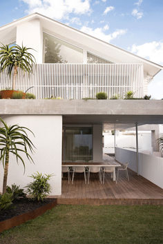 a white house with wooden decking and plants on the top floor, next to an outdoor dining area