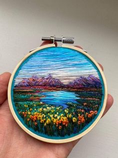 a hand holding a small embroidery art piece with mountains in the background and flowers on it