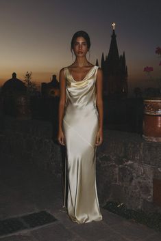 a woman in a long white dress standing on a stone wall at night with the sun setting behind her