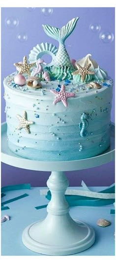 there is a blue cake with stars and mermaid decorations on the top, sitting on a white pedestal