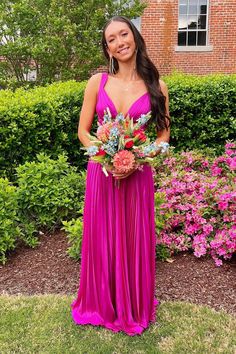 a woman standing in front of some bushes holding a flower bouquet and wearing a pink dress