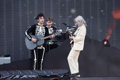 two men in skeleton suits playing guitars on stage with an older man standing next to them