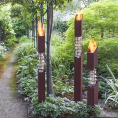 three metal sculptures in the middle of a garden with trees and bushes around them, lit by torches