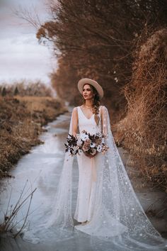 a woman in a wedding dress and hat standing on a frozen path with her bouquet