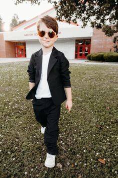 a young boy wearing sunglasses and a black suit in front of a building with trees