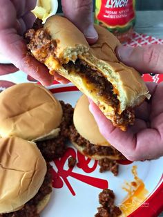 two hands holding up a sandwich with cheese and chili on it next to some hamburgers