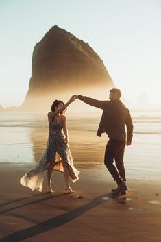 a couple dancing on the beach in front of a rock formation at sunset, with one holding the other's hand