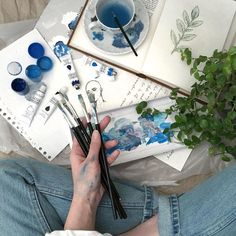 a person sitting on the floor with paint and brushes in their hands next to some plants