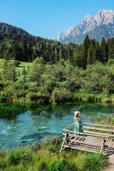 a woman standing on a wooden bridge over a river with mountains in the back ground