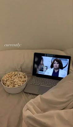 a laptop computer sitting on top of a bed next to a bowl of cereal and a bowl of popcorn