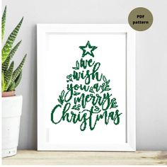 a cross stitch christmas tree with the words merry and stars in green on it, next to a potted plant