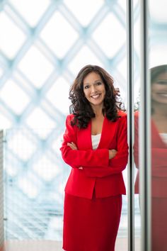 a woman in a red suit standing next to a tall glass wall with her arms crossed