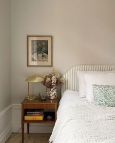 a white bed sitting next to a wooden table with flowers on it and a painting above the bed