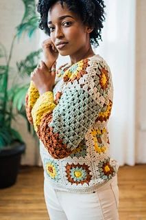 a woman standing in front of a plant wearing a crocheted sweater and white pants