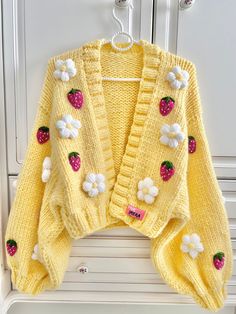 a yellow sweater with white flowers and strawberries on the front is hanging from a hook