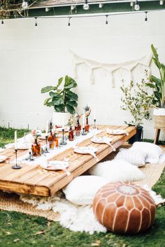 a picnic table set up with food and drinks on it in the grass next to a white brick wall