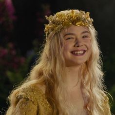 a woman with long blonde hair wearing a gold crown and smiling at the camera while standing in front of trees