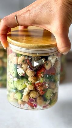 a hand holding a jar filled with vegetables and chickpeas, on top of a counter