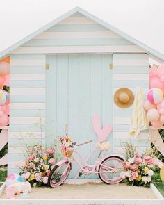 a pink bicycle parked in front of a beach hut with balloons and flowers around it