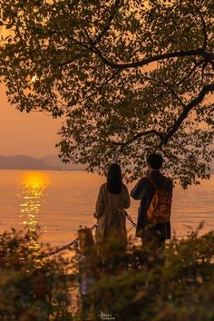 two people standing next to each other on a lake shore at sunset with the sun in the distance