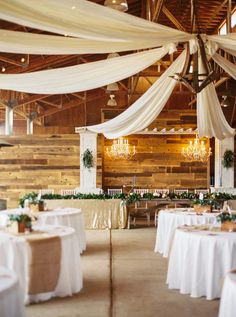 the inside of a barn decorated with white linens, greenery and chandeliers